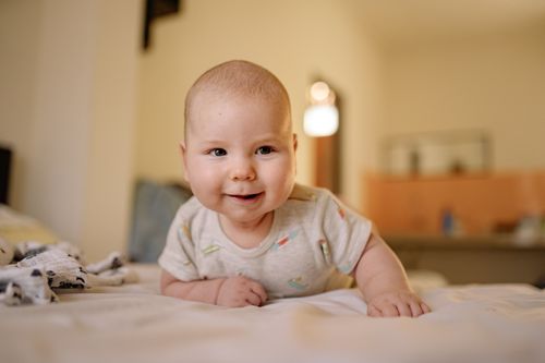 Free Cute baby in a onesie smiling while lying on a bed indoors. Stock Photo