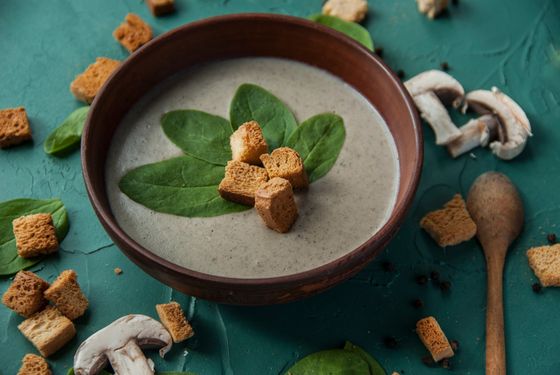 Pale creamy mushroom soup topped with spinach leaves and rye croutons, mushrooms scattered nearby