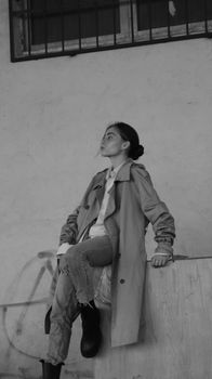 Chic woman in coat and jeans posing against urban backdrop in black and white.
