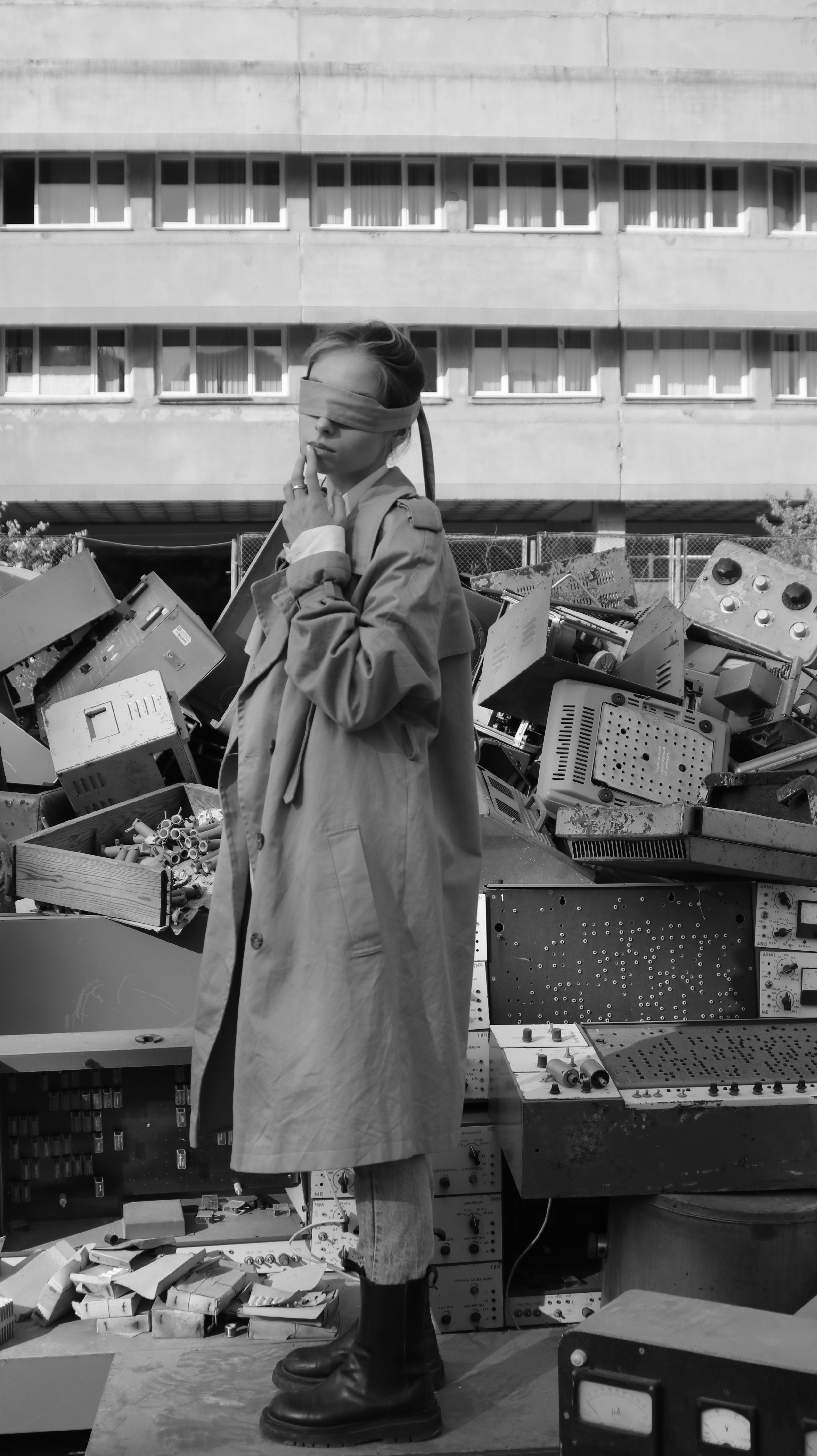 A fashion model in a coat and boots poses blindfolded among electronic debris, creating a striking monochrome image.