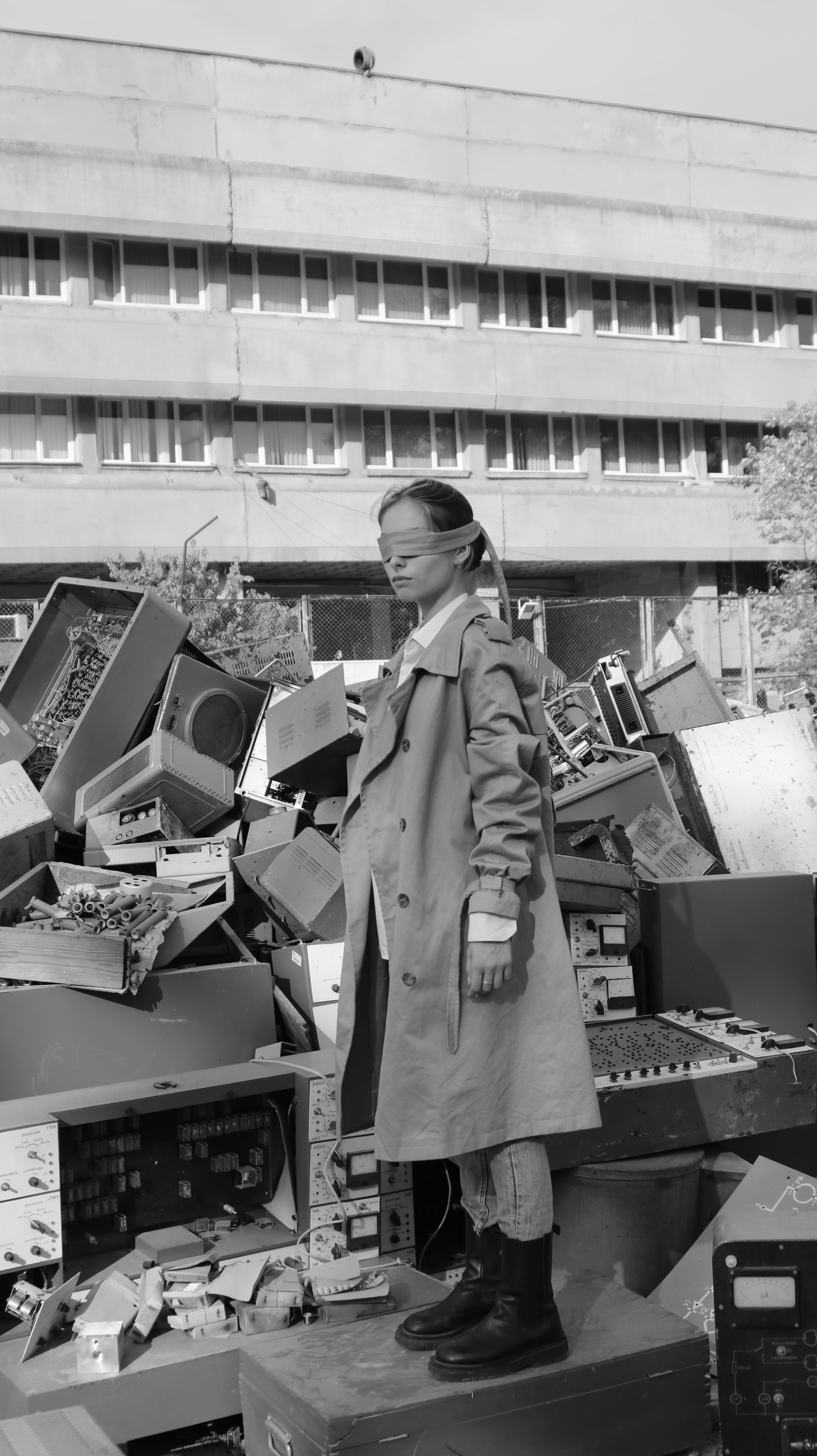 Black and white photo of a blindfolded woman standing amidst electronic waste in an urban setting.