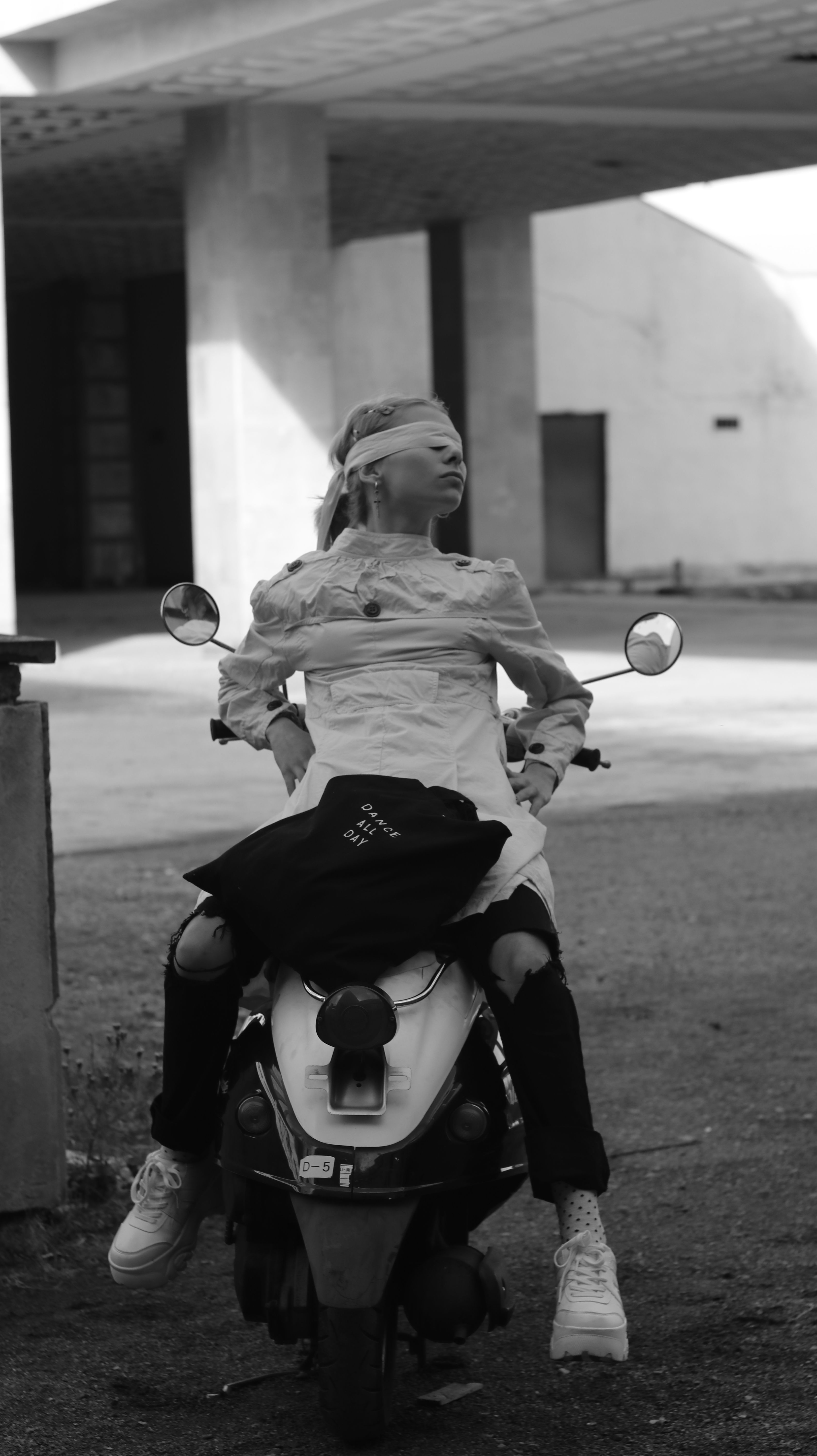 Monochrome image of a fashionable woman in ripped jeans sitting blindfolded on a scooter.