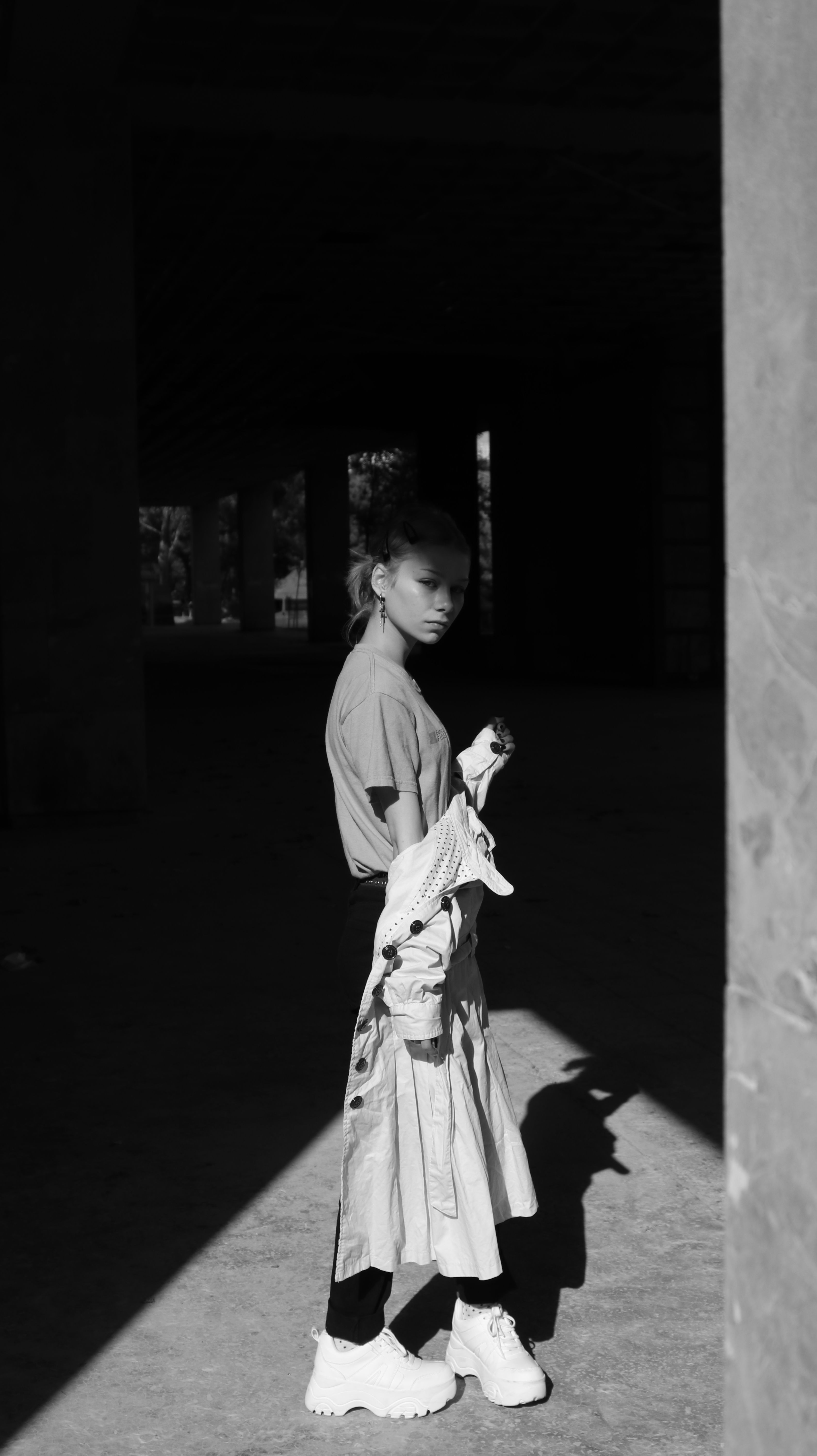A dramatic black and white portrait of a woman standing in sunlight under a building.