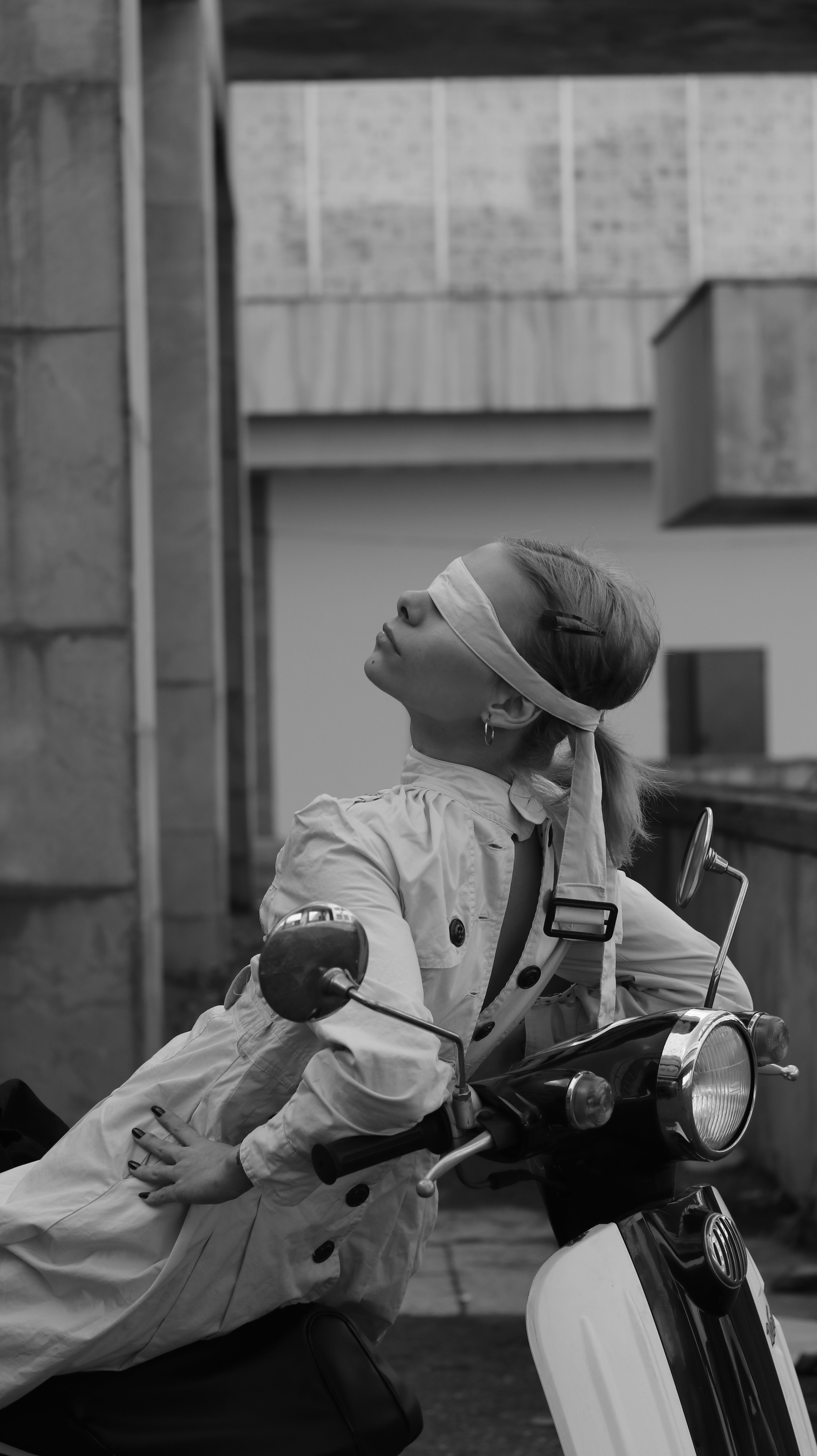 Artistic black and white photo of a stylish woman blindfolded on a scooter, conveying fashion and intrigue.