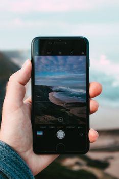 Hand holding a smartphone capturing a scenic beach view with ocean waves.