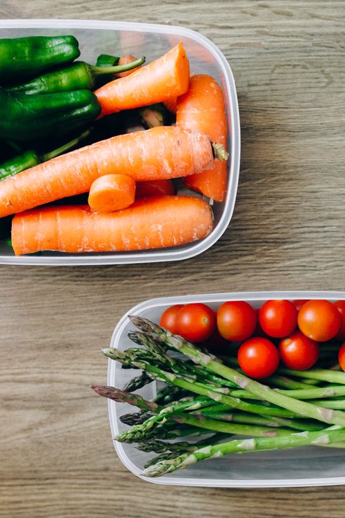 Free Flat lay of vibrant vegetables including carrots, tomatoes, and asparagus in plastic containers. Stock Photo