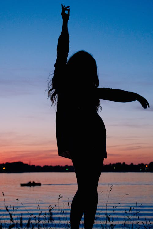 Silhouette of anonymous female with hand up standing on lake coast at bright sundown