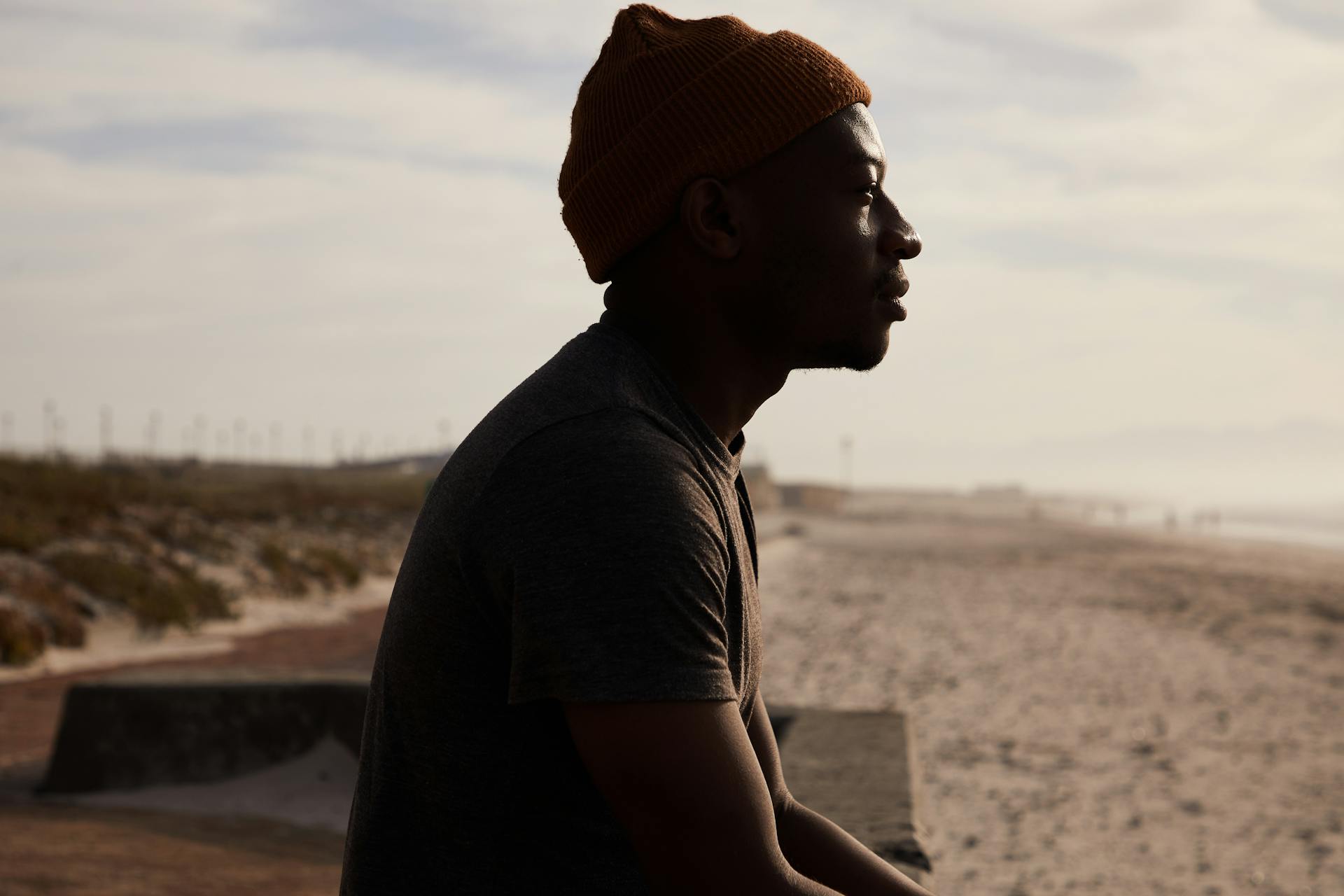 Side view of African American male in hat sitting on cement border on sandy shore near sea against cloudy sky