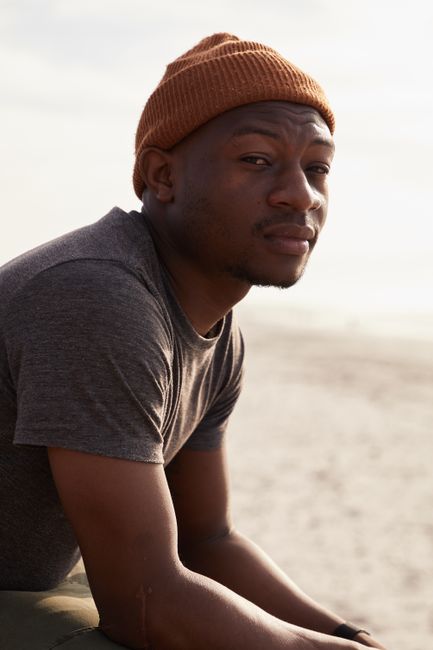 Pensive African American male wearing hat looking at camera while sitting on sandy seaside against cloudless sky on blurred background