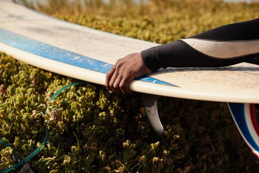 Crop unrecognizable ethnic male athlete in diving suit with aged surfboard near lush green bushes in summer