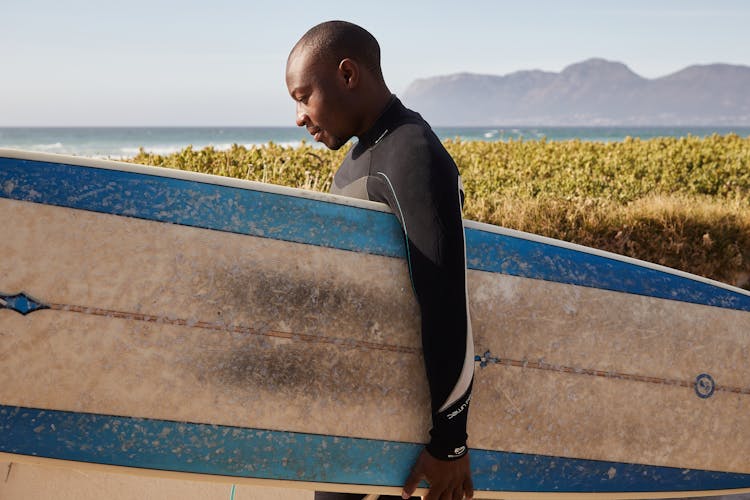 Black Athlete With Old Surfboard Near Shrubs On Beach