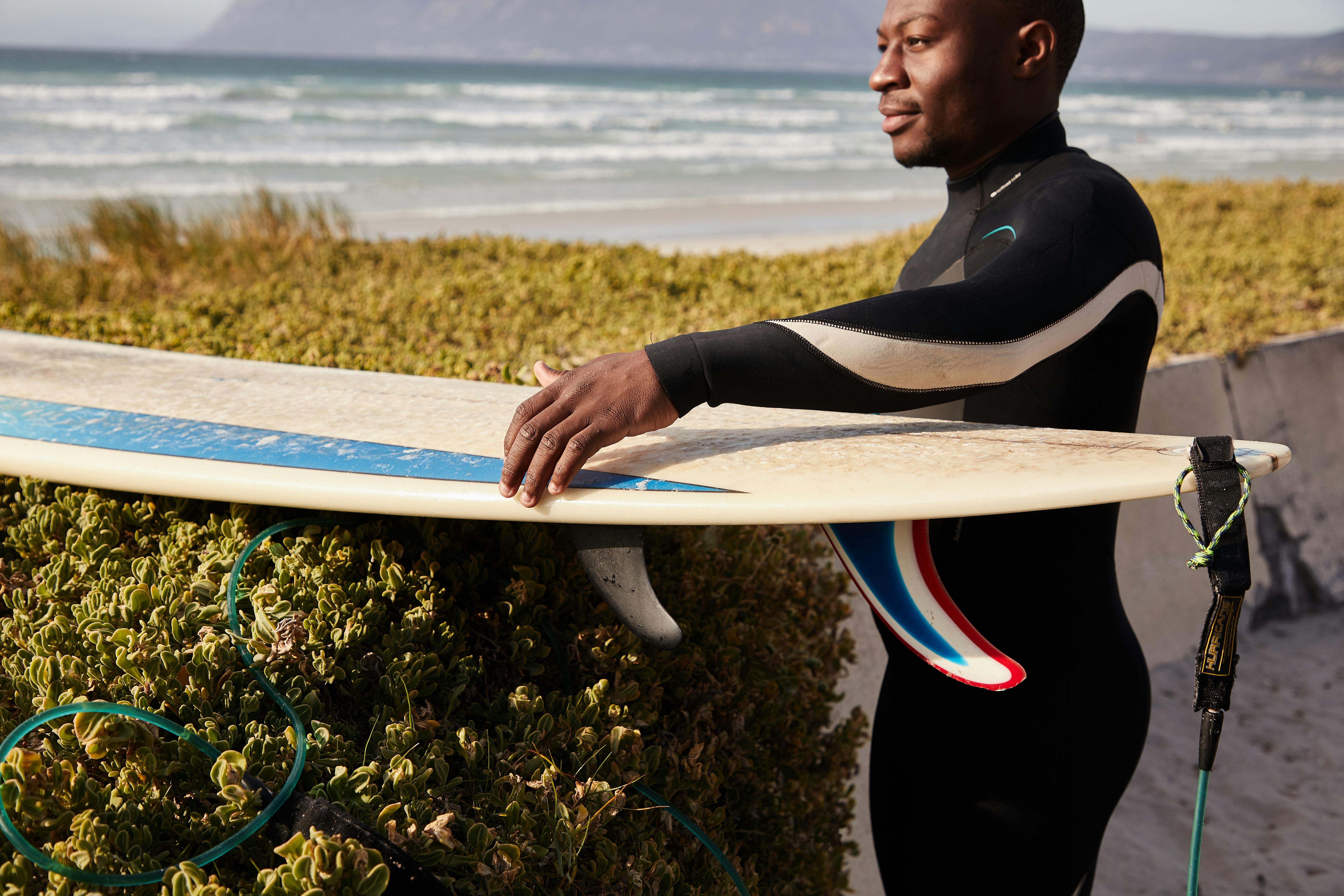 Side view of crop young contemplative African American athlete in diving suit with surfboard near greenery shrubs looking forward on sea coast