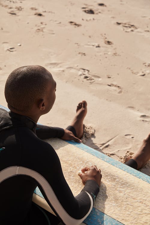 From above of African American traveler in wetsuit looking away while chilling on wet sandy coast covered with footprints in sunlight