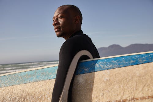 Pensive black man with board for surfing on coast of sea