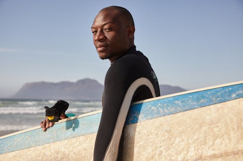 Side view of African American professional surfer looking at camera and carrying surfboard on blurred background of rocky coast