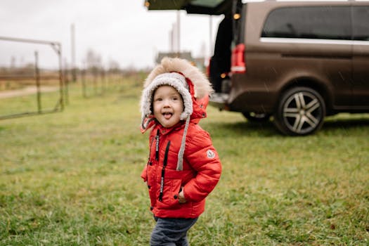 Young child in red puffer jacket enjoying a rainy day outdoors, standing near a vehicle.