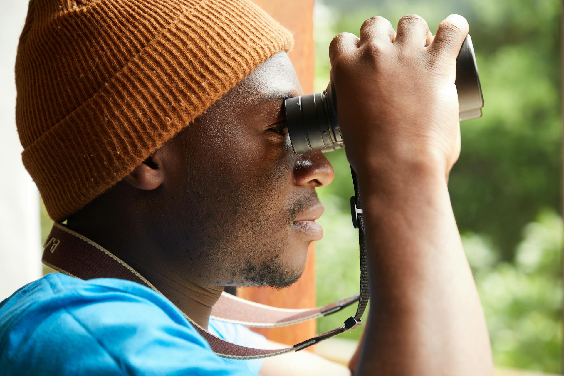 Side view of anonymous African American male in trendy hat looking through binoculars while standing against opened window