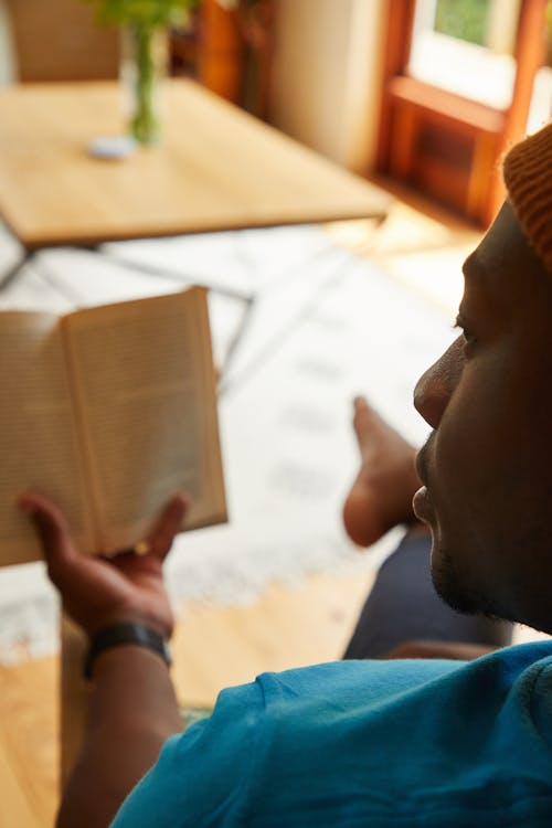 Free Crop African American male sitting on couch and reading book while spending weekend at home Stock Photo