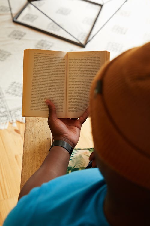 Free From above of crop faceless African American male in trendy hat sitting at table and reading book Stock Photo