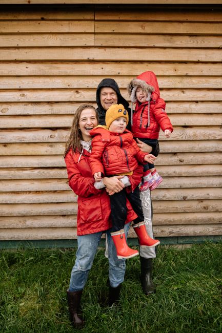 Family of four in matching red jackets smiling outdoors in front of wooden wall.