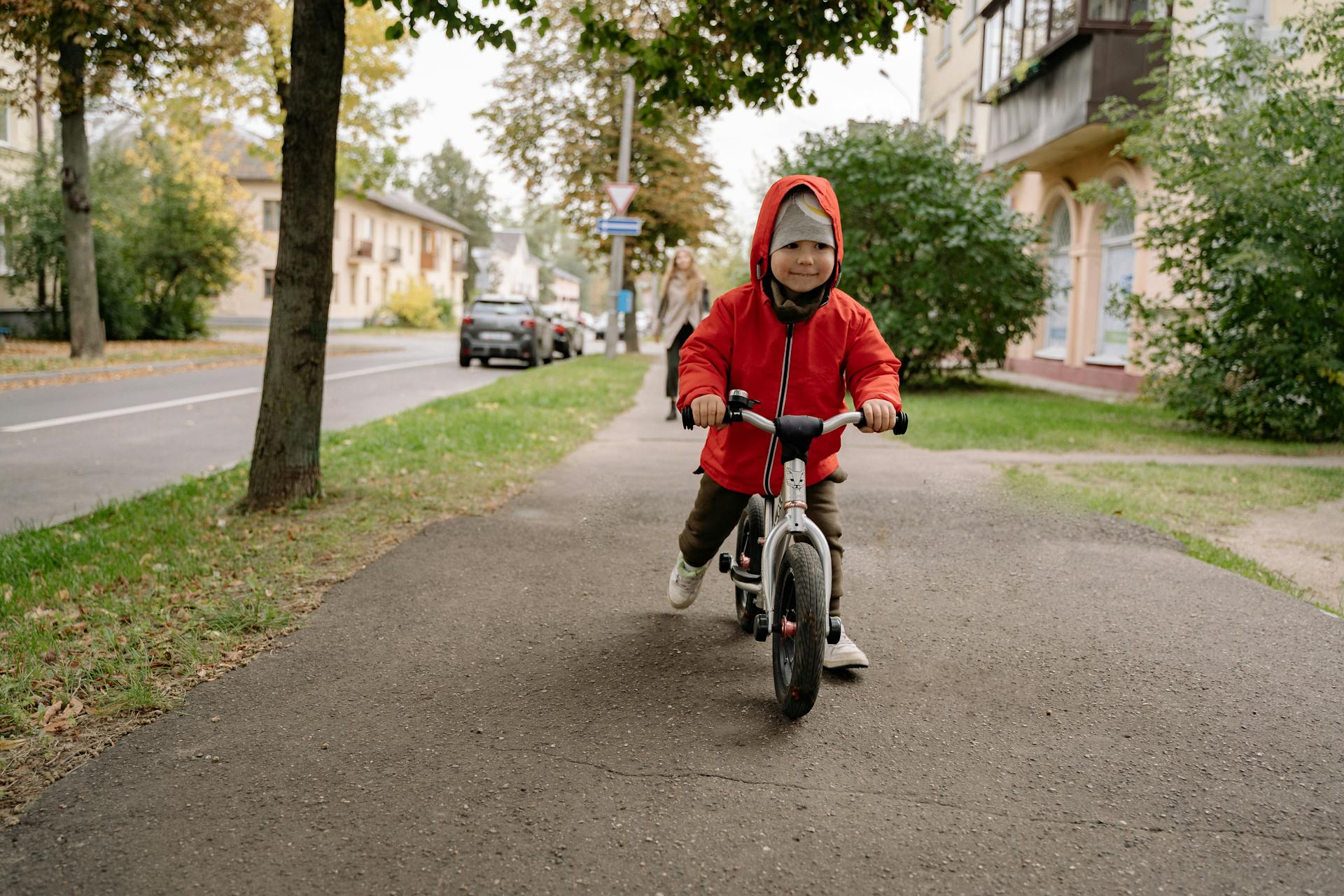 Child in a red jacket riding a balance bike on a tree-lined urban sidewalk. Autumn setting.