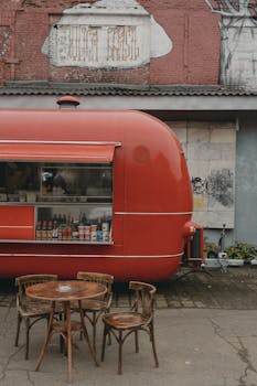 Red food truck parked by a brick wall, rustic outdoor seating setup.