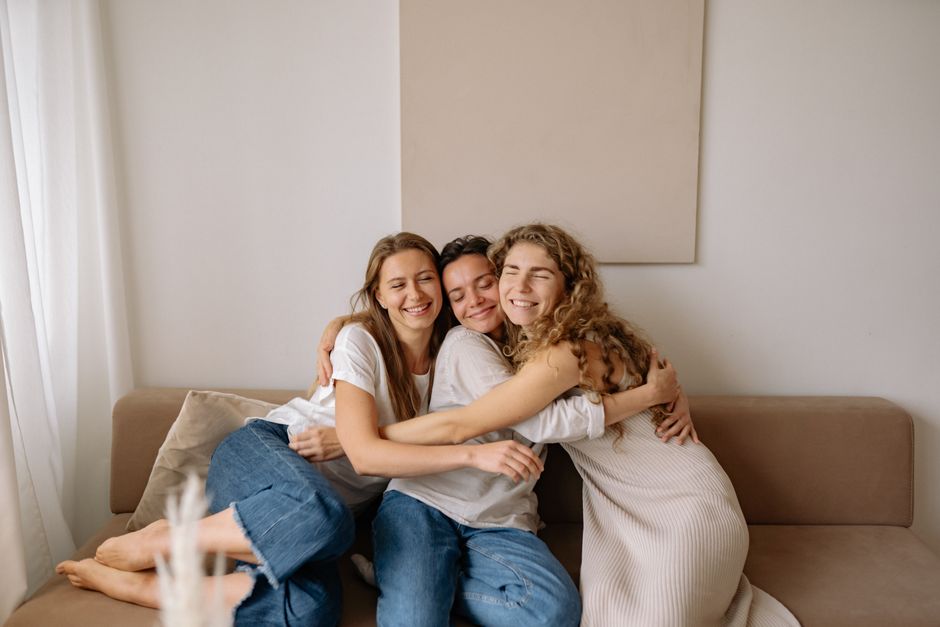 Fotomural Tres amigas disfrutan de un abrazo sincero en un sofá acogedor, irradiando calidez y cercanía.