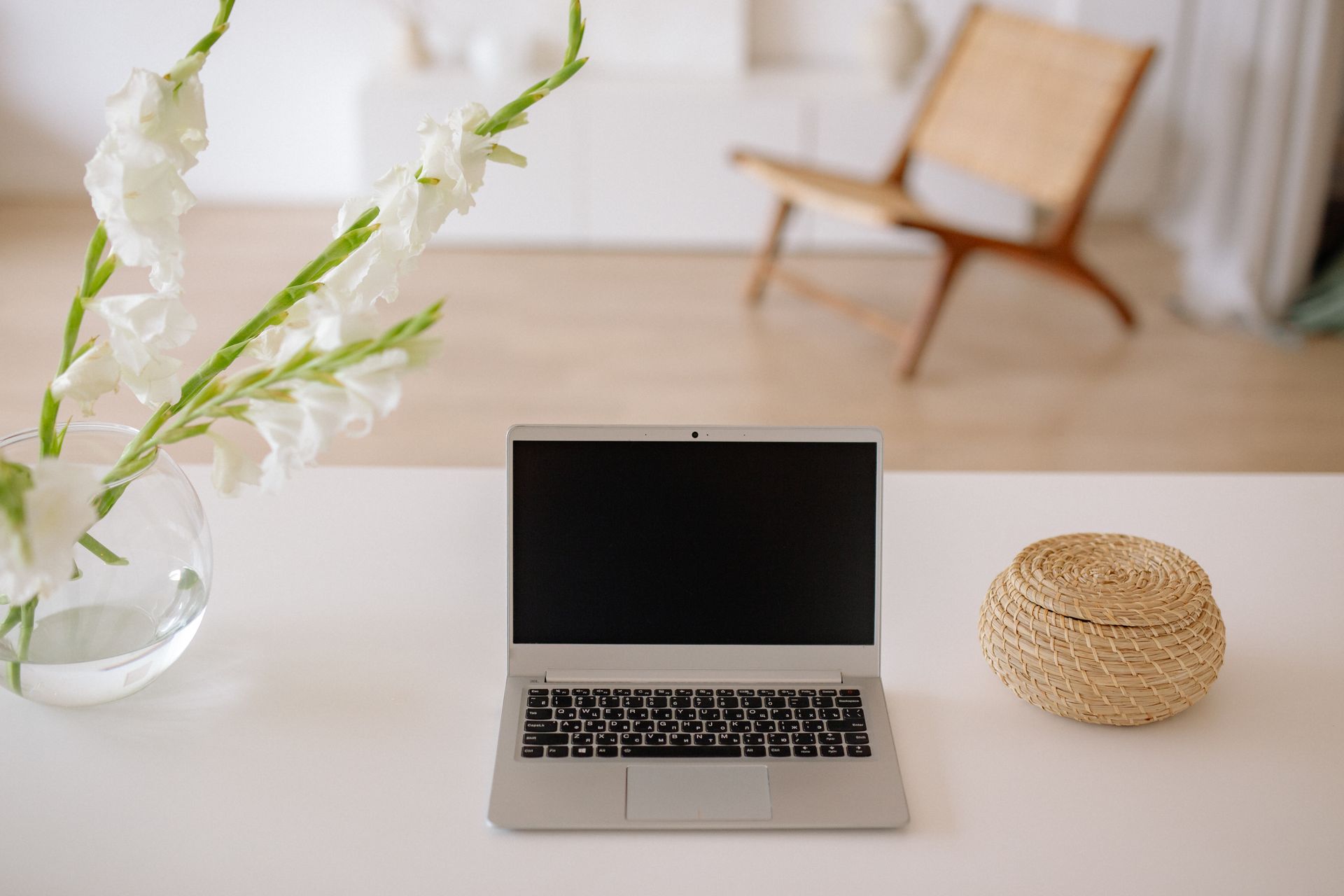 A serene home office space featuring a laptop, white flowers, and wicker basket on a desk.