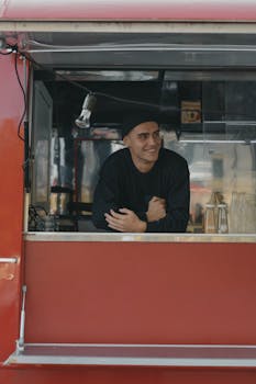 A young man in a beanie serving at a red food truck, representing modern street food culture.