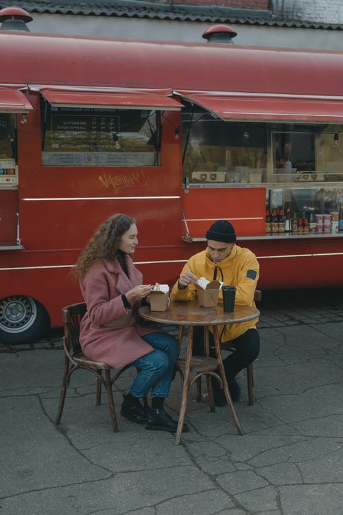 Free A couple enjoys takeout food at a table beside a red food truck outdoors. Stock Photo