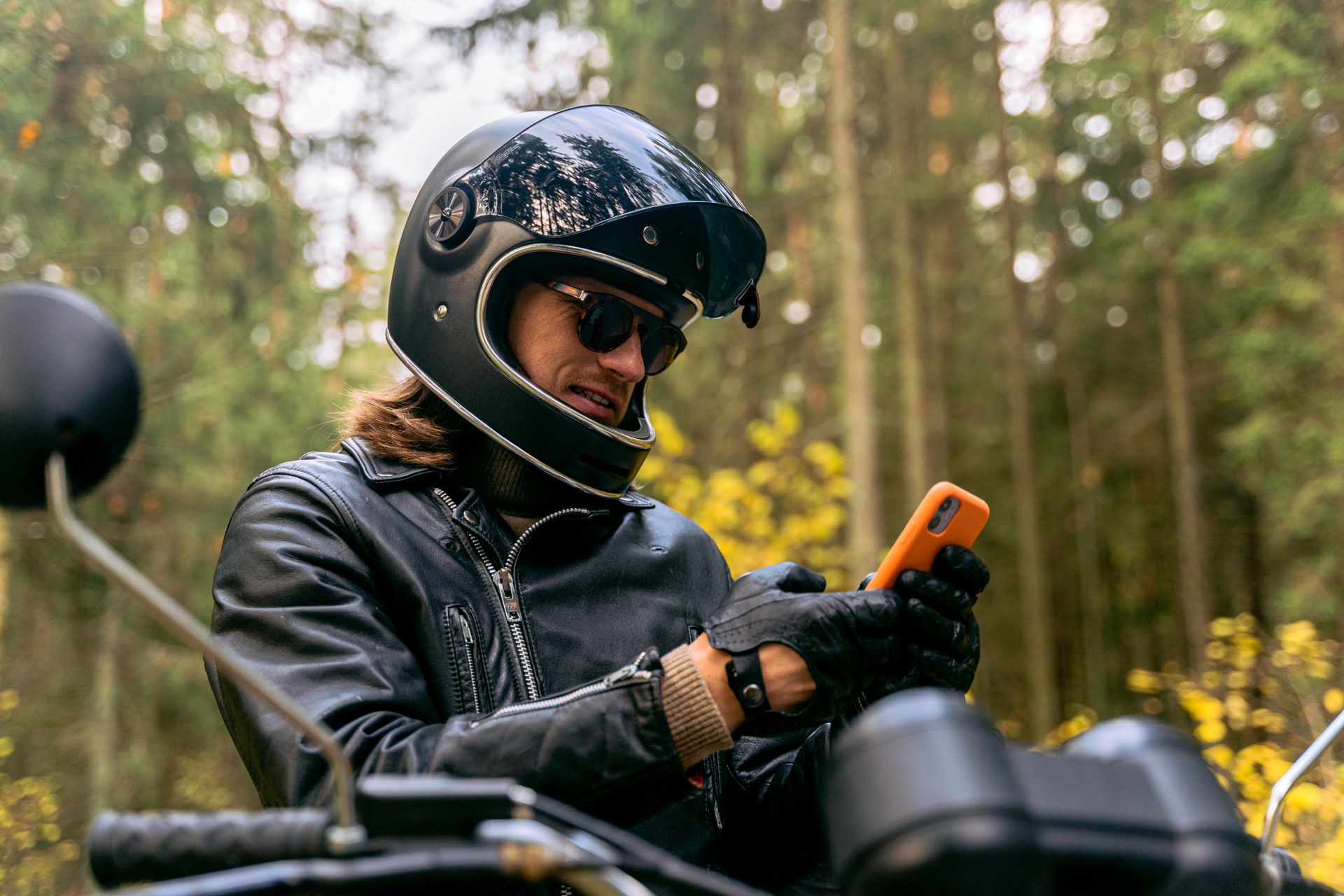 A biker wearing protective gear checks his smartphone while parked in a forest.