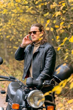 Man in black leather jacket on a motorcycle making a phone call amidst autumn forest.