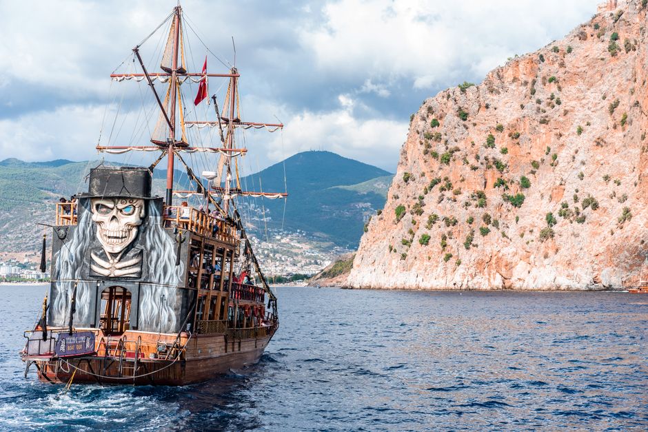 Wooden pirate boat with sailing mast and painted skull floating on rippling sea near rocky mountain against grassy hilly area