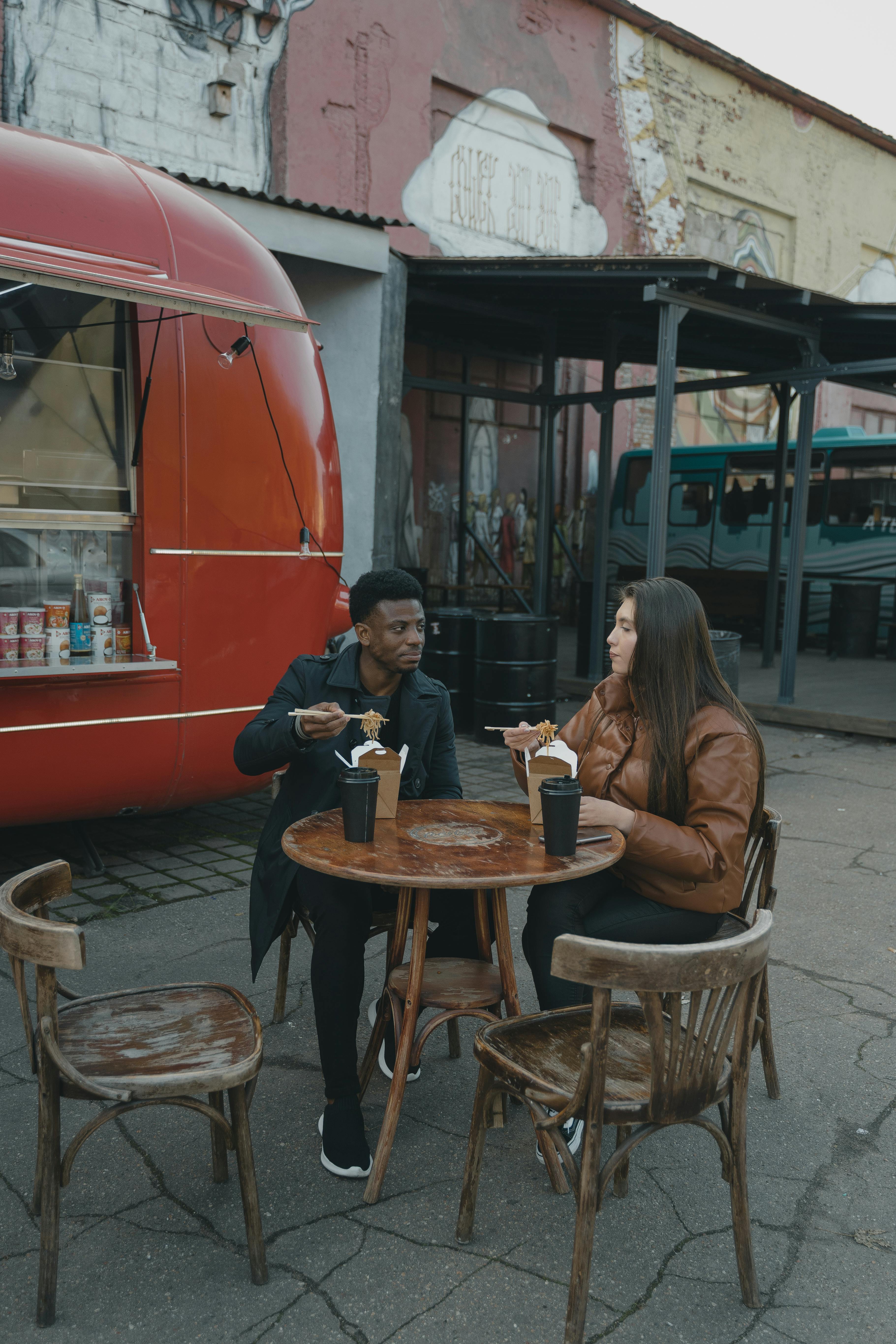 Interracial couple enjoying street food outside a vibrant red food truck on a cool day.