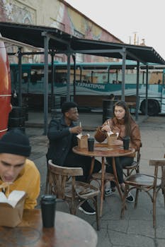 Young adults enjoying takeaway food at an outdoor street food corner with colorful bus backdrop.
