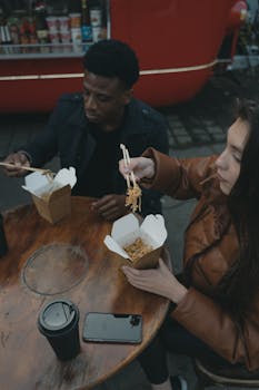 A couple enjoys takeout noodles at a food truck with outdoor seating.