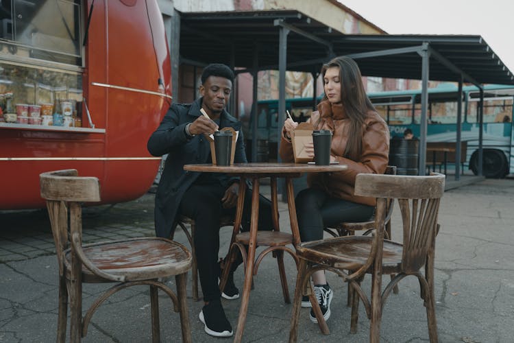 A Man In A Black Coat And A Woman In A Brown Jacket Dining Outdoors