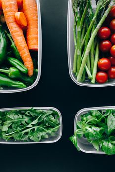 Top view of assorted fresh vegetables in plastic containers on a black background.