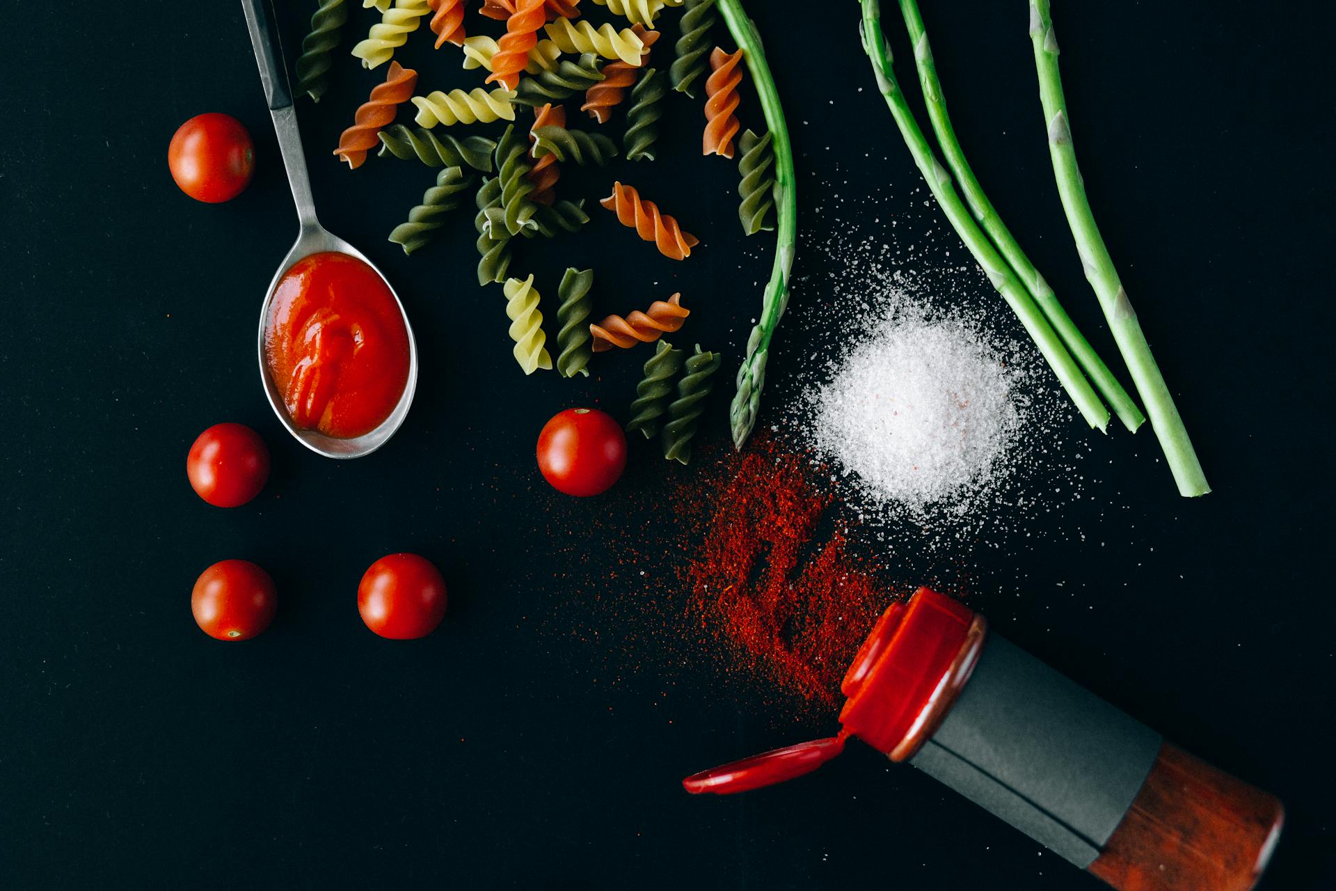 Flatlay of fresh Italian ingredients including pasta, tomatoes, and spices on a dark blue background.