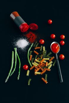 Colorful uncooked fusilli, tomatoes, asparagus, and spices on a dark surface.