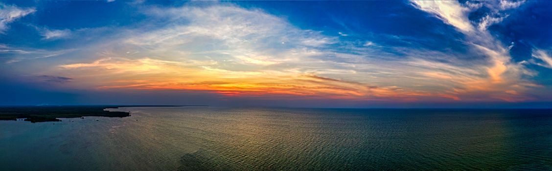 Panoramic view of wavy sea with horizon and island under bright sky with clouds at sundown