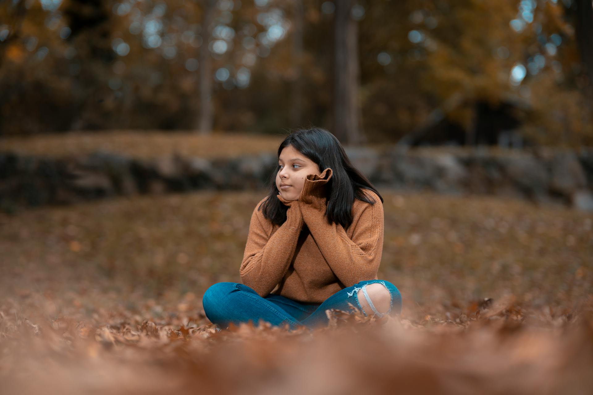 A woman sits on fallen leaves wearing a brown sweater in Glen Cove, capturing the essence of fall.