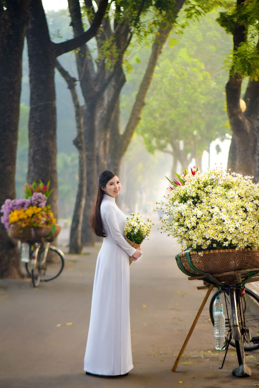 Elegant Vietnamese woman in traditional attire holding flowers beside bicycles on a picturesque stre