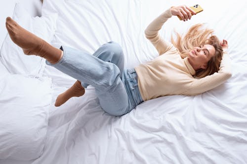 A Woman in Beige Turtle Neck Long Sleeves Looking at a Mobile Phone while Lying on the Bed