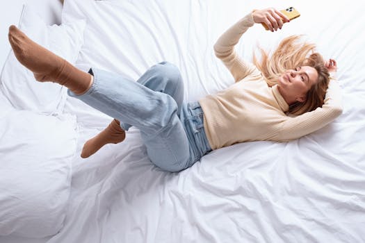 Young woman lying on bed using smartphone, dressed in casual jeans and sweater, enjoying modern technology.