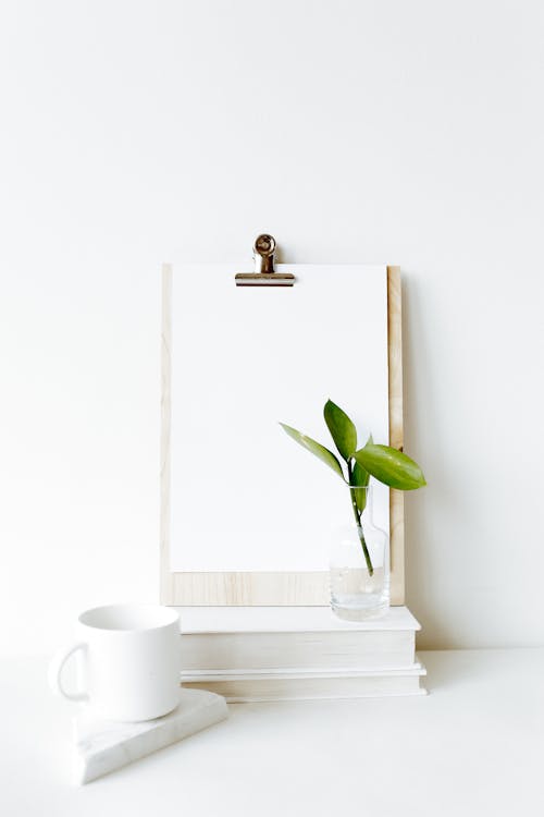 Free Composition of clipboard with empty sheet placed on stacked books near white mug and green plant leaf Stock Photo