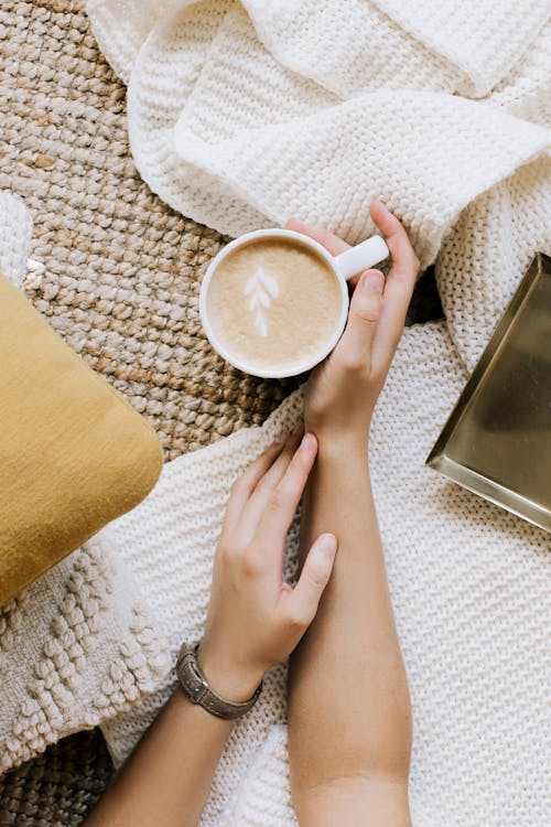 Top view crop unrecognizable female resting hands with cup of freshly brewed cappuccino on cozy soft cloth