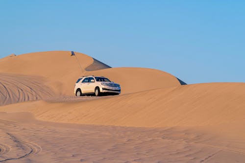 Low angle of modern off road car with flag on pole driving along sandy dunes against cloudless blue sky in desert
