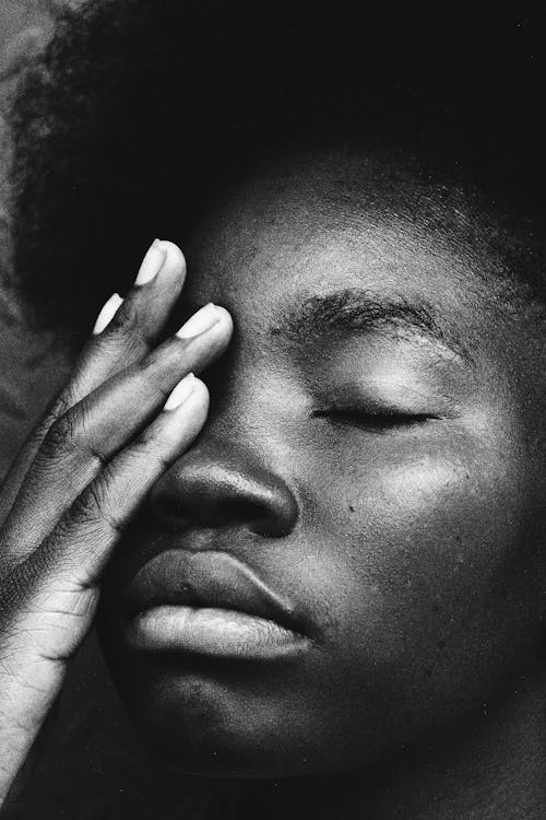 Black and white crop sad African American female with Afro hairstyle standing with eyes closed and touching face in thoughts
