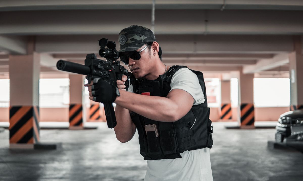 A man dressed in tactical gear aims a rifle in a shaded parking area, suggesting a training scenario.